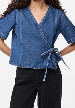 Femme portant un top cache-cœur en denim bleu à manches courtes noué sur le côté avec un pantalon noir sur fond blanc.