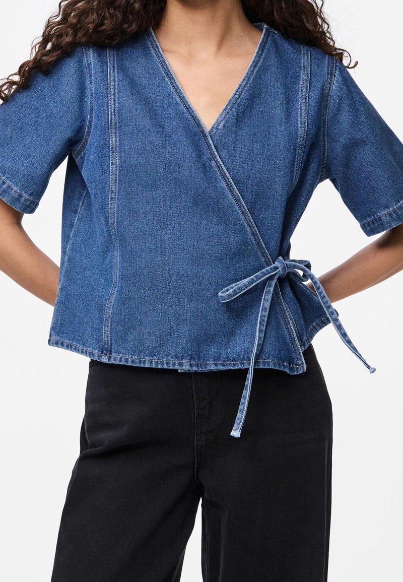 Femme portant un top cache-cœur en denim bleu à manches courtes noué sur le côté avec un pantalon noir sur fond blanc.