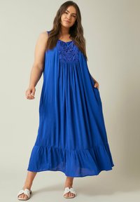 Blaues Maxikleid mit ärmellosem Design, aufwendigem besticktem Oberteil, geraffter Taille und gestuftem Volantsaum. Kombiniert mit weißen Sandalen.