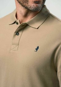 Beige polo särk, mis on valmistatud tekstureeritud kangast, millel on nööpide rida ja väike tume sinine kudumärgis vasakul rinnaesisel.