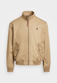 Beige bomberjack gemaakt van lichtgewicht stof, met een opstaande kraag, ritssluiting, twee zijzakken en een klein logo op de borst.