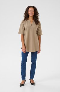 Beige, kortärmad tunika med V-ringning och avslappnad passform. Kombineras med blå skinny jeans och svarta högklackade skor med en rosettdetalj.