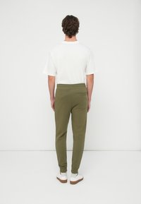 Muž v bielom tričku, olivovo zelených teplákoch s vreckom na zadnej strane a bielych teniskách, stojaci čelom k jednoliatemu svetlému pozadiu.