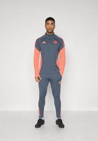Atletski trenirku u sivoj boji s koraljnim akcentima. Sadrži jaknu sa četvrtastim zatvaračem, duge rukave i sužene hlače. Prisutna je Adidasova oznaka.