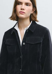 Giacca di velluto a coste scura con colletto strutturato, tasche sul petto e dettagli con bottoni argento. Indossata sopra una camicia bianca, presenta una superficie testurizzata.