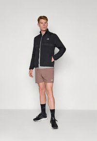 Black lightweight jacket with a zip front, grey t-shirt underneath, brown shorts with logo, black sneakers, standing pose against a plain background.