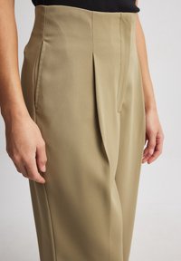 Béžové kalhoty s vysokým pasem, s záhyby vpředu a hladkou, texturovanou tkaninou. Na bocích jsou viditelné kapsy u švů.