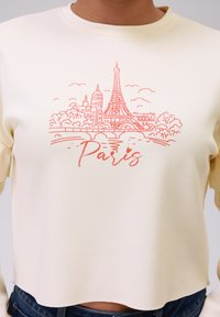 Felpa corta color crema con una grafica arancione della Torre Eiffel e il testo "Paris", caratterizzata da una superficie in tessuto morbido.