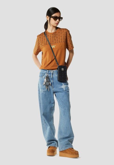 Vrouw draagt een bruin patroon blouse, blauwe wijde jeans met patches, bruine plateau schoenen, zwarte crossbody tas, zonnebril en een sleutelhanger accessoire.