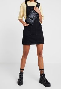 Person som bär en beige oversized t-shirt, svart denim overallklänning, svart axelväska, nätstrumpor och kraftiga svarta platåskor.