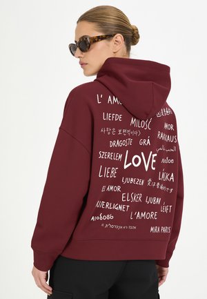 Frau mit Dutt und Schildpatt-Sonnenbrille trägt einen bordeauxfarbenen Hoodie mit dem Wort "love" in mehreren Sprachen auf dem Rücken.