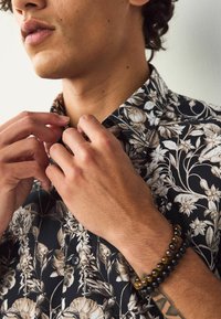 Hemd mit Blumenmuster in Schwarz und Beige. Kurzärmelig, mit spitzem Kragen und Knopfverschluss. Perlenarmbänder zieren die Handgelenke.