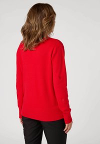 Maglione rosso lavorato a maglia con orlo e polsini a coste; presenta ritagli a forma di cuore sulle maniche; morbido al tatto, vestibilità rilassata. Indossato con pantaloni neri.