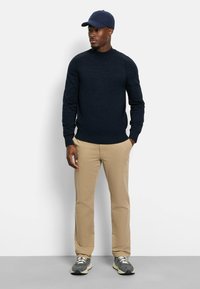 Marineblauer Rundhals-Pullover, khakifarbene Hose, grau-weiße Sneaker und eine passende marineblaue Mütze. Der Pullover zeichnet sich durch ein strukturiertes Strickdesign aus.