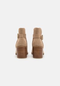 Une paire de bottes chevilles en daim beige avec des talons en bois empilés et des sangles à boucle sur le côté, présentée de l'arrière sur un fond blanc.