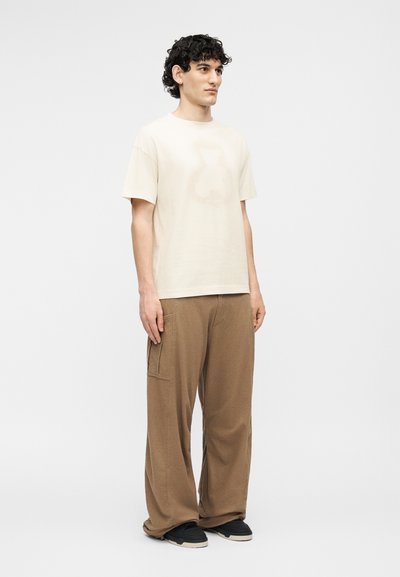 Νέος άνδρας με σκουρόχρωμα σγουρά μαλλιά, ντυμένος με απλό μπεζ κοντομάνικο μπλουζάκι, φαρδιές καφέ παντελόνες με πλαϊνές τσέπες και μαύρα αθλητικά παπούτσια.