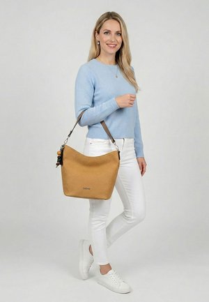 Mujer rubia con suéter azul claro y pantalones blancos lleva un gran bolso marrón texturizado con llavero de cuentas, de pie frente a un fondo liso.