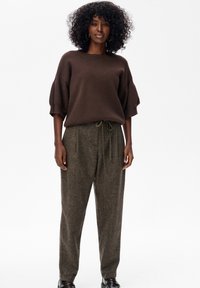 Vrouw die staat en een losse bruine top met ellebooglange mouwen, hoogsluitende joggingsbroek van wol en donkere instappers draagt.