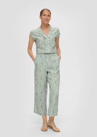 Groene jumpsuit met een geometrisch wit patroon, korte mouwen, v-hals, elastische tailleband en zijzakken. Gecombineerd met platte sandalen.