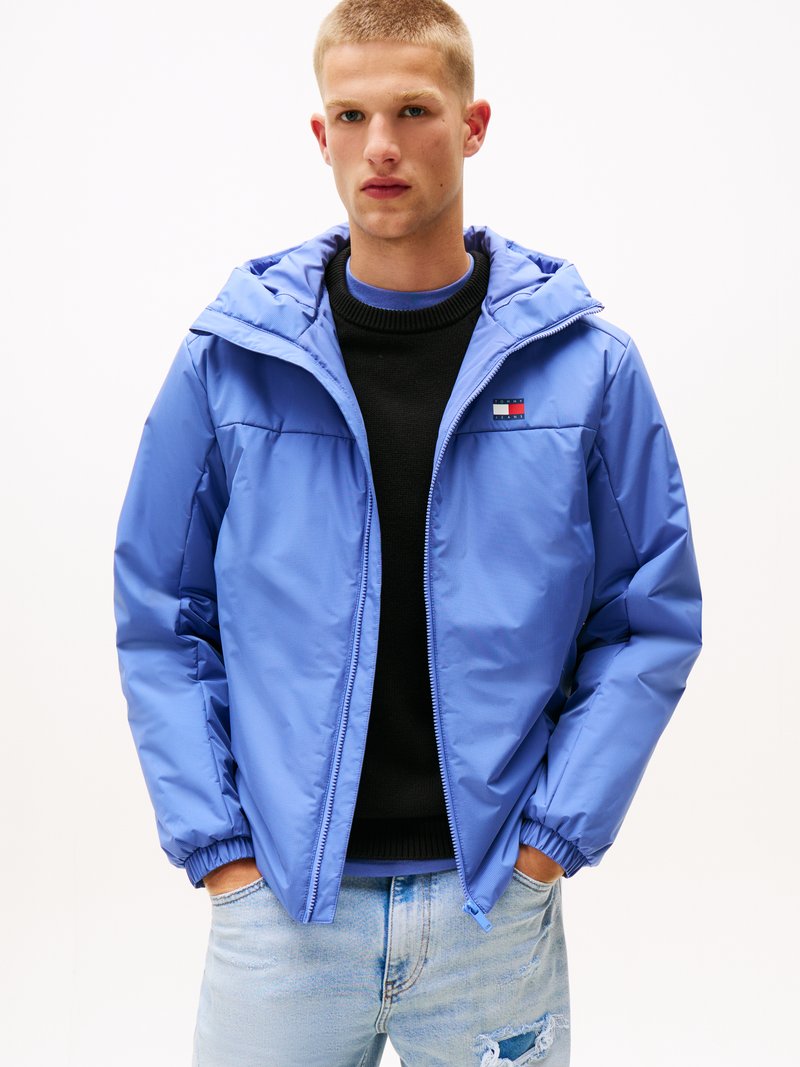 Giacca a vento blu chiaro in materiale liscio, con zip frontale, polsini elasticizzati e una patch con logo sul petto. Indossata sopra un maglione nero.