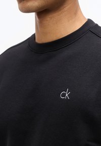 Άνδρας που φοράει μαύρο φούτερ με στρογγυλή λαιμόκοψη και μικρό λευκό λογότυπο "ck" κεντημένο στο αριστερό στήθος.