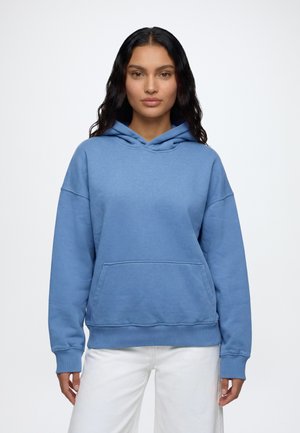Femme aux longs cheveux foncés portant un sweat à capuche bleu clair et un pantalon blanc, debout devant un fond blanc uni.