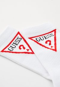 Chaussettes en coton blanches avec un logo triangulaire rouge comportant le texte "GUESS" et un point d'interrogation, mettant en avant une texture côtelée et des détails colorés.