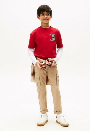 Rotes T-Shirt mit kurzen Ärmeln und schwarzem "H"-Logo, darunter ein weißes Langarmshirt, beige Cargo-Hosen und weiße Sneakers mit beige-farbenen Sohlen.