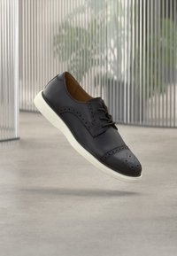 Chaussure derby en cuir noir avec détails perforés, bout rond et semelle en caoutchouc blanche. Dotée d'un laçage traditionnel et d'une texture lisse.