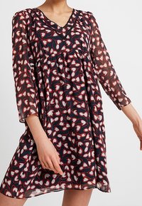 Valkoinen sydänkuvioinen, läpikuultava tummansininen mekko, jossa on V-pääntie, kolme neljäsosapituista hihaa ja kootut vyötärö. Yksityiskohtainen pääntien viimeistely.