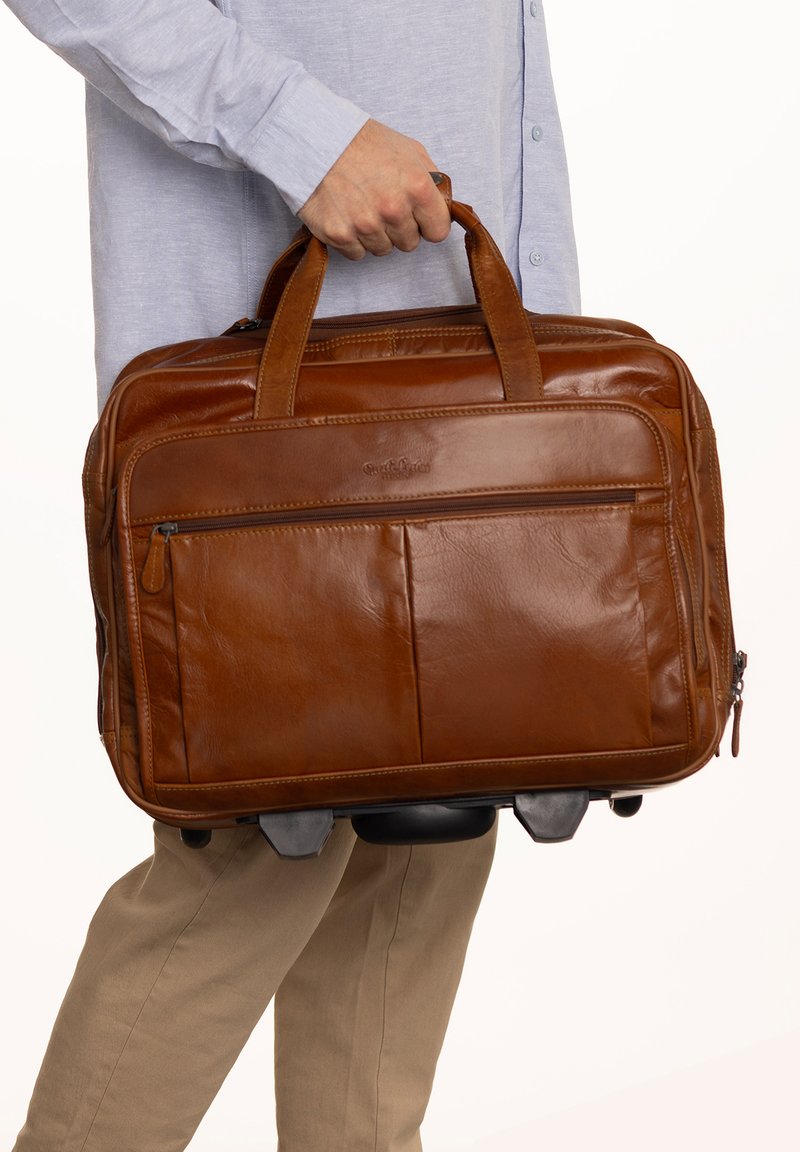 Man in light blue shirt and beige pants holding brown leather briefcase with wheels against white background.
