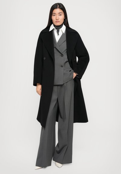 Double-breasted black wool coat over a gray three-piece suit with large pants, white shirt, and pointed white heels. Smooth textures.