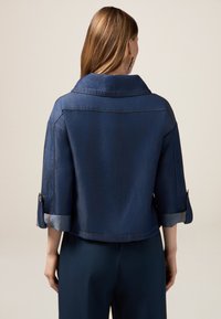 Chaqueta de mezclilla en azul oscuro con un corte ajustado, cuello ampliado y mangas remangadas, con puños en un azul claro en contraste y detalles de costura.