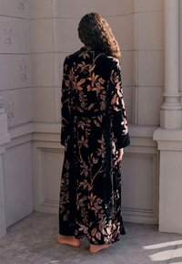 Ilgas juodas aksominis chalatas su dideliais rožiniais gėlių raštais ir diržu, plačiomis rankovėmis ir minkšta tekstūra, dėvimas basomis pėdomis.