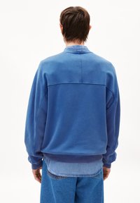 Blaues Sweatshirt mit Rundhalsausschnitt und übergroßer Passform, gerippten Bündchen und Saum, mit einer genähten Nahtdetails über dem Rücken. Über einem Denimhemd getragen.