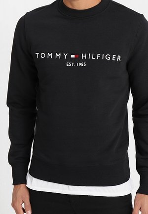 Sort sweatshirt med lange ærmer med teksten "Tommy Hilfiger", rød og hvid flaglogo og "EST. 1985" trykt på brystet.