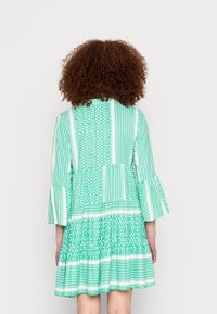 Vestido verde con patrón y mangas acampanadas, que presenta diversos diseños geométricos, volantes en la cintura y un corte a la altura de la rodilla.