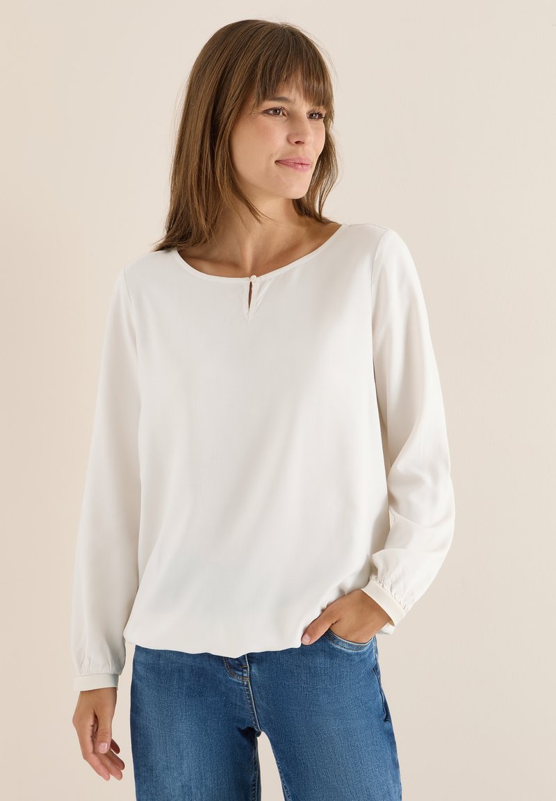 Blusa bianca a maniche lunghe con scollatura rotonda, caratterizzata da un piccolo dettaglio a chiave e polsini arricciati, abbinata a jeans blu.