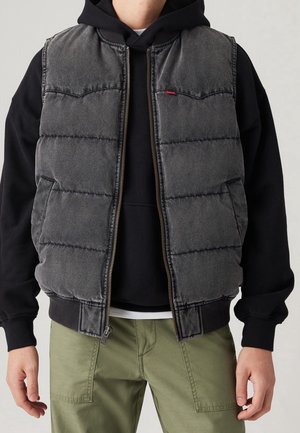 Homme portant un sweat à capuche noir, un gilet matelassé gris zippé et un pantalon vert, montré du cou aux hanches sur un fond uni.