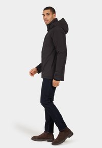 Črna, lahka, toplotno izolirana jakna z zadrgo in kapuco, v kombinaciji z temnimi jeans hlačami in rjavimi nubuk škornji. Preprost dizajn, brez vzorcev.