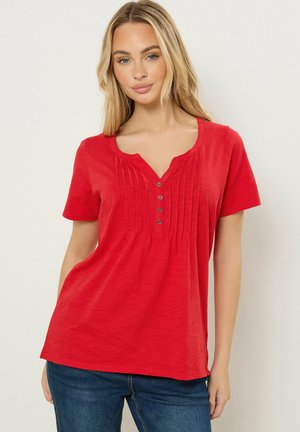 Frau trägt ein rotes Kurzarmoberteil mit Knöpfen vorne und Falten, kombiniert mit blauen Jeans, vor einem schlichten hellen Hintergrund stehend.