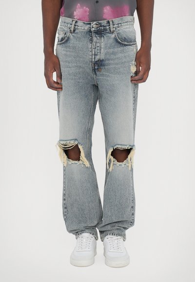 Jean en denim délavé avec de grandes déchirures aux genoux, coupe décontractée et bords effilochés. Associé à des baskets blanches sur un fond neutre.