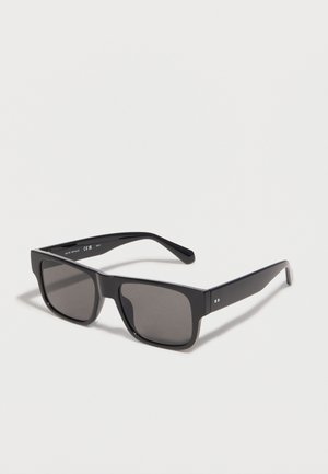 Μαύρα ορθογώνια γυαλιά ηλίου με σκουρόχρωμους φακούς και χοντρές πλευρικές βάσεις, τοποθετημένα σε λευκή επιφάνεια και στραμμένα προς τα δεξιά.