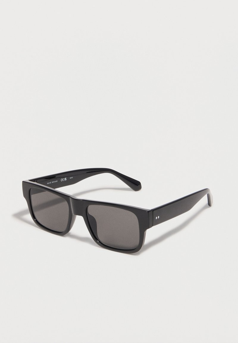 Μαύρα ορθογώνια γυαλιά ηλίου με σκουρόχρωμους φακούς και χοντρές πλευρικές βάσεις, τοποθετημένα σε λευκή επιφάνεια και στραμμένα προς τα δεξιά.