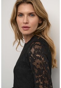 Svart spetsblus med långa ärmar, som har intrikata blommönster, texturerat tyg och en figurnära design. Slät, helsvart bod.