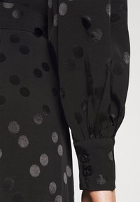 Détail d'une robe noire avec des manches bouffantes, ornée de pois en velours noirs et gris de différentes tailles, et de poignets à boutons. Texture de tissu lisse.