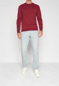 Pull col rond bordeaux associé à un jean bleu clair et des baskets blanches. Le pull présente un petit logo sur la poitrine.