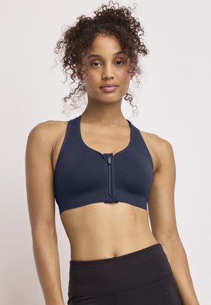 Young woman with curly hair wearing a navy zip-front sports bra and black high-waisted leggings, standing against a plain light background.