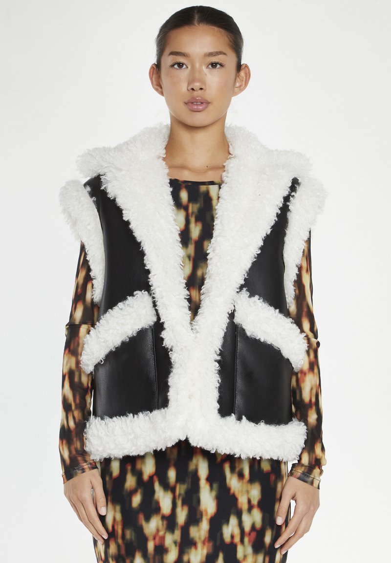 Gilet in pelle nera con colletto e bordo in pelliccia bianca, dotato di due tasche frontali e design aperto. Lo sfondo è bianco uniforme.