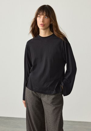 Kobieta z długimi brązowymi włosami, ubrana w czarną bluzkę z długim rękawem i szare szerokie spodnie, stojąca na tle jednolitego jasnego tła.